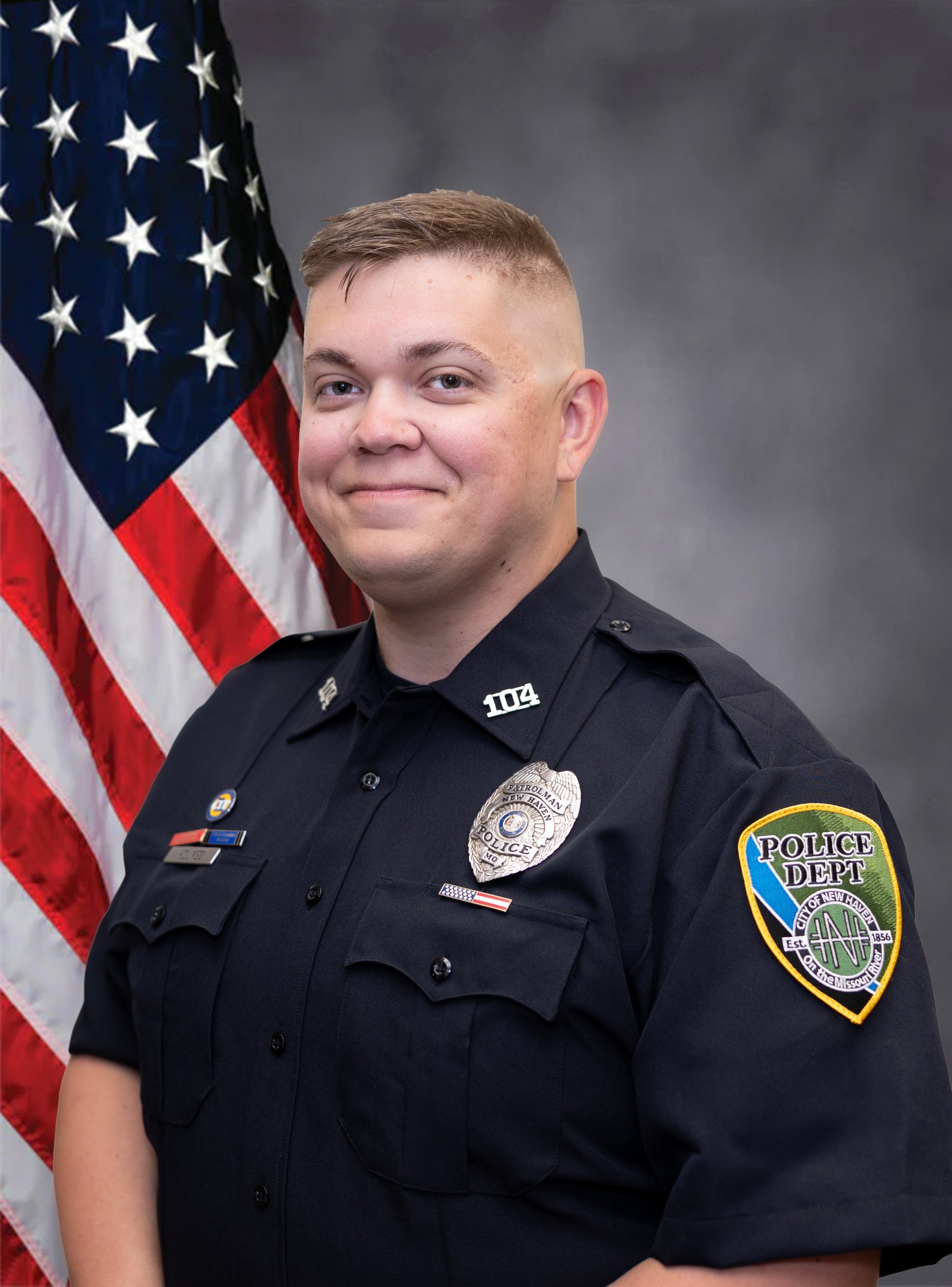 Uniformed police officer portrait with U.S. flag behind; blue “Police Dept” patch on chest.